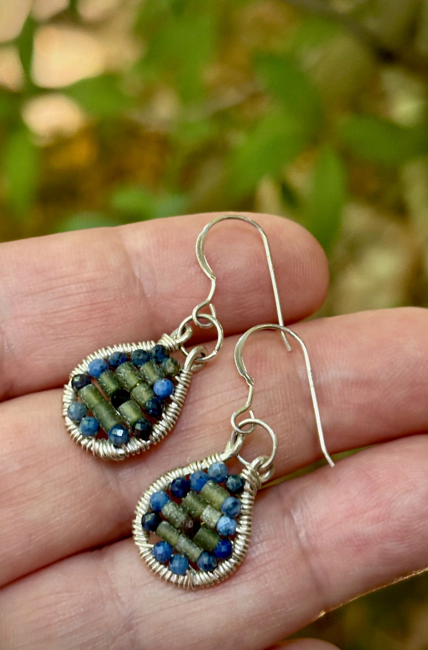 Teardrop-shaped wire-wrapped earrings with jade and garnet stones on a hand against a blurred natural background
