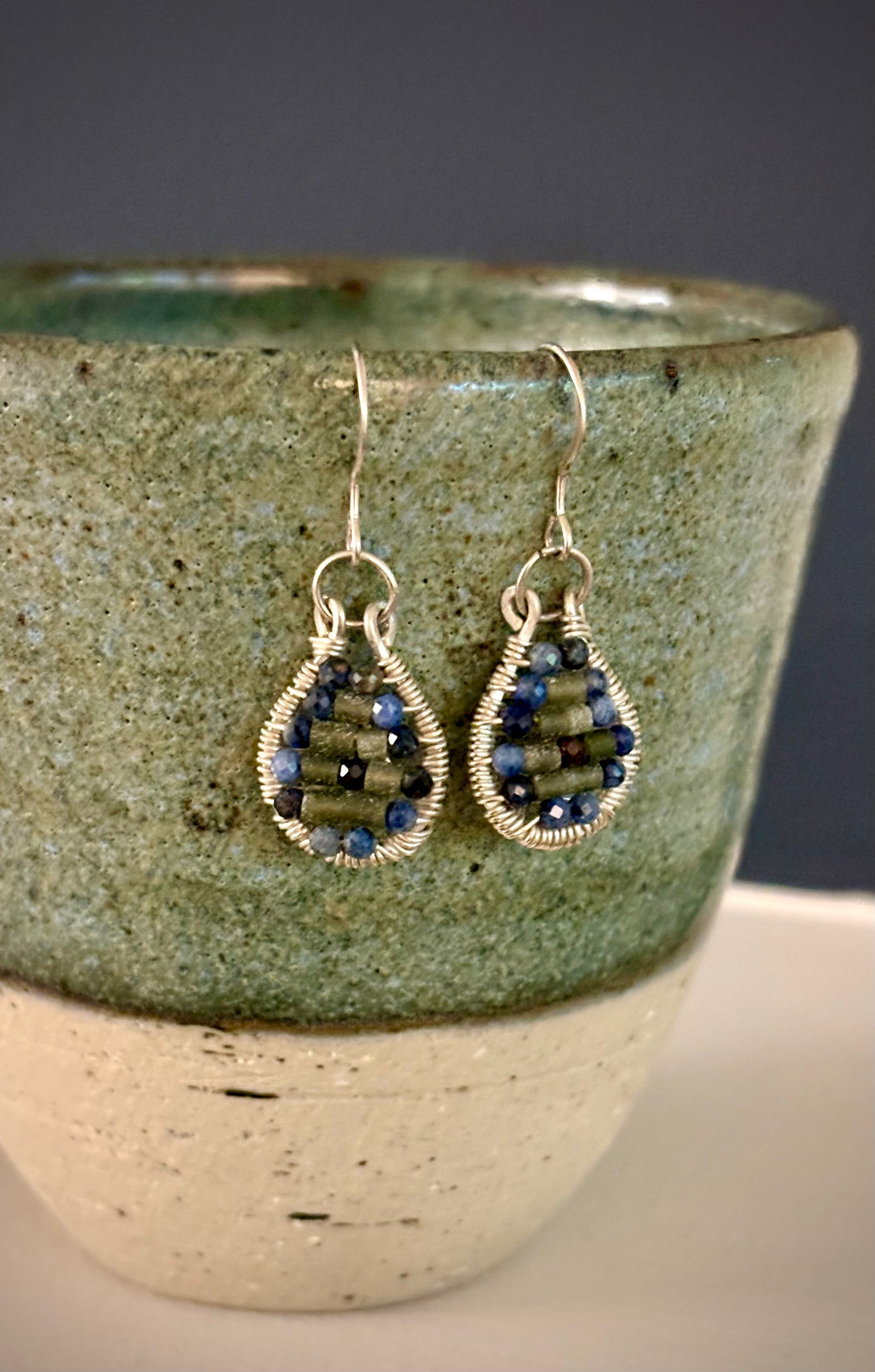 Teardrop-shaped wire-wrapped earrings with garnet and jade beads hanging on a textured ceramic cup.