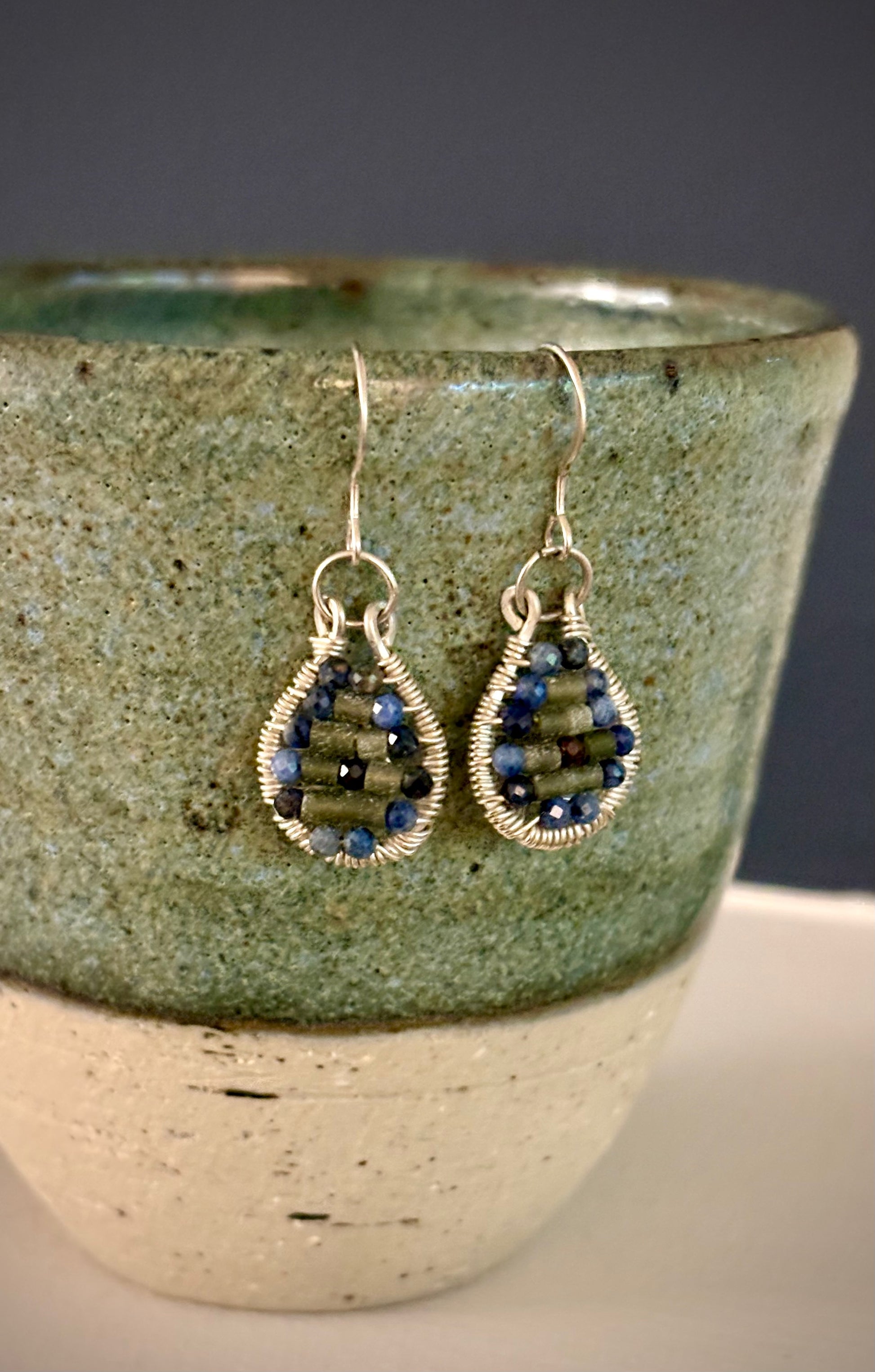 Teardrop-shaped wire-wrapped earrings with garnet and jade beads hanging on a textured ceramic cup.