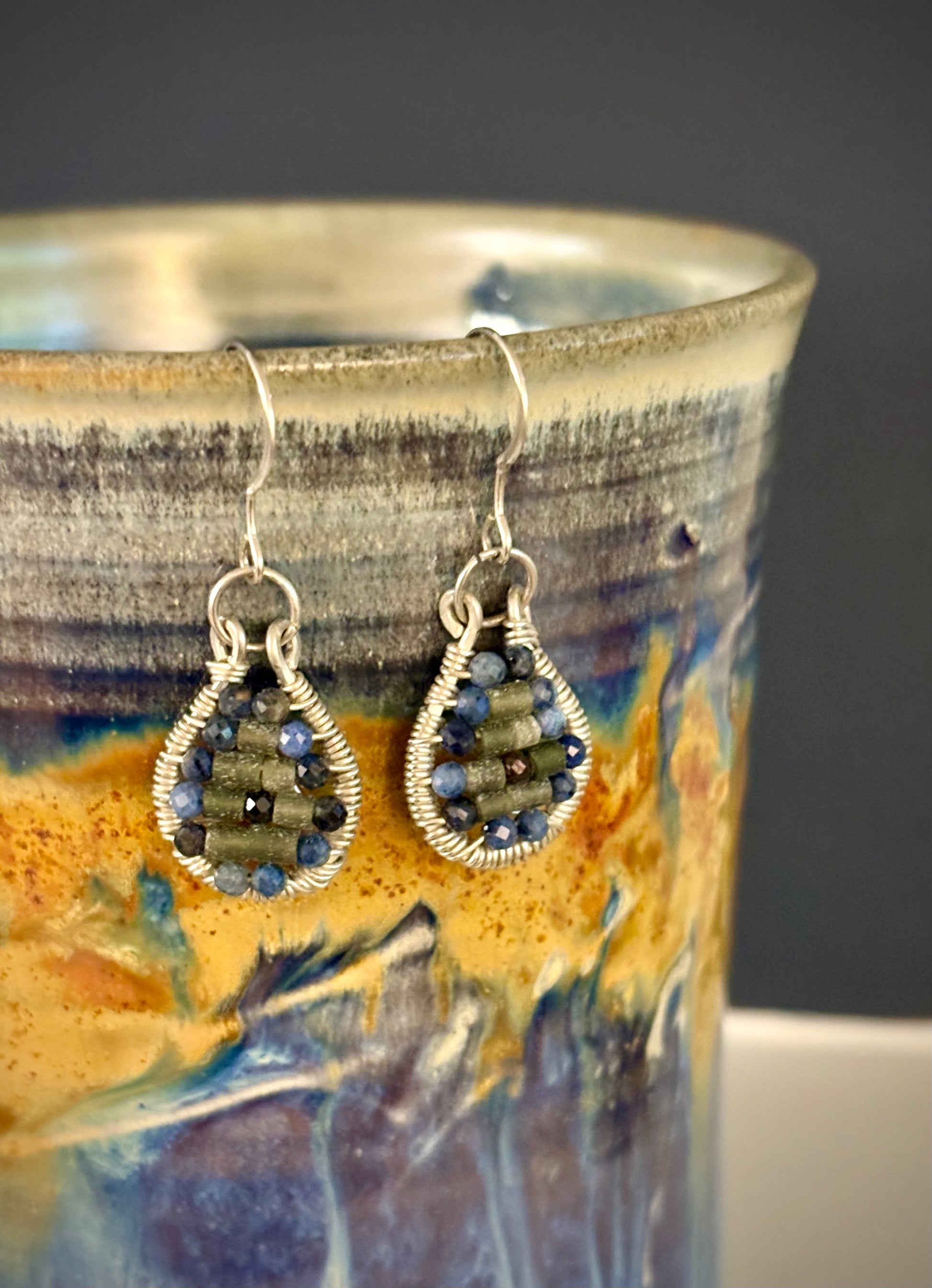 Pair of silver wire-wrapped earrings with garnet and jade stones on a ceramic mug with a colorful design.